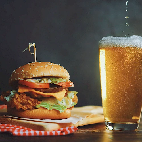 A juicy fried chicken burger with lettuce and tomato beside a frosty glass of beer, on a rustic wooden table.