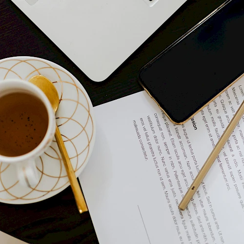 A coffee cup on a saucer with a wooden stirrer, beside a smartphone, scissors, laptop corner, and papers with a pencil on a dark desk.