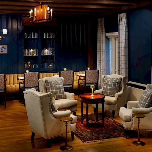 An elegant lounge with upholstered chairs, a glowing table lamp, tall windows with curtains, and a bookshelf in a cozy blue room.