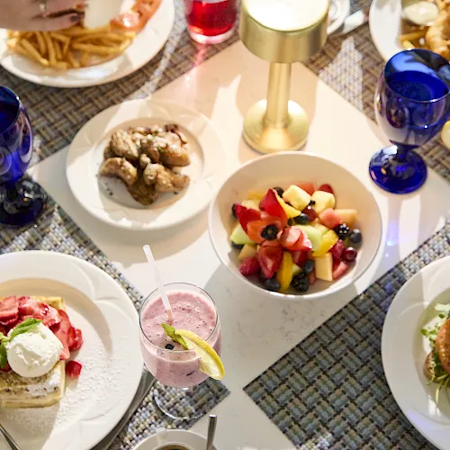 A dining table set with fries, fresh fruit salad, pastries, and pink/blue drinks in glasses, ready to eat.