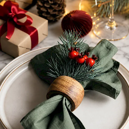 A holiday table setting with a green napkin, pine branches, red baubles, a pinecone, wrapped gifts, and warm string lights.