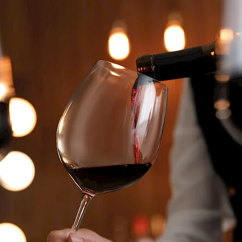 A bottle pours red wine into a stemmed glass, with warm bokeh lights glowing in the background.