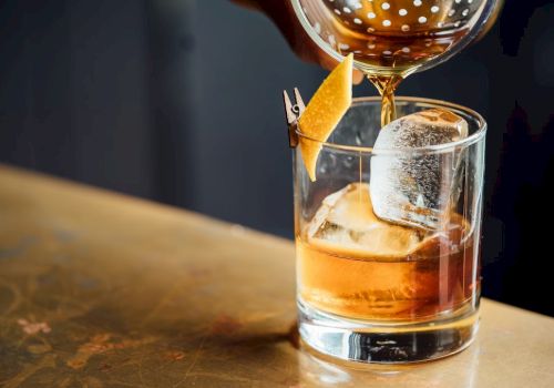 A glass of whiskey on the rocks being poured with a garnish orange twist, captured at a bar counter.