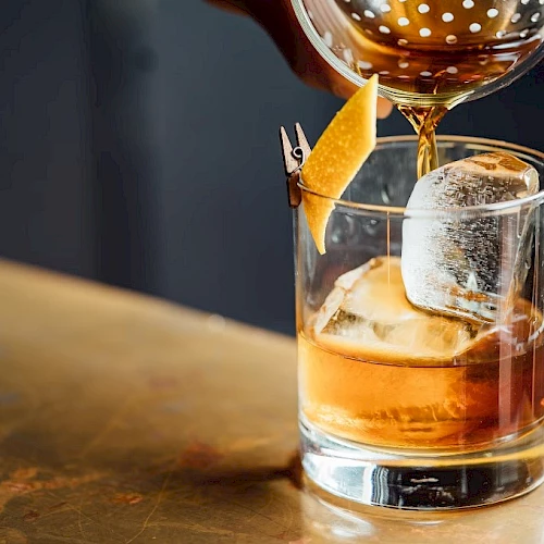 A glass of whiskey on the rocks being poured with a garnish orange twist, captured at a bar counter.