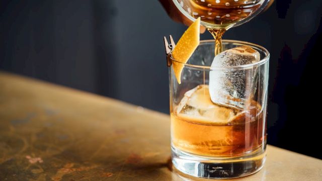 A glass of whiskey on the rocks being poured with a garnish orange twist, captured at a bar counter.