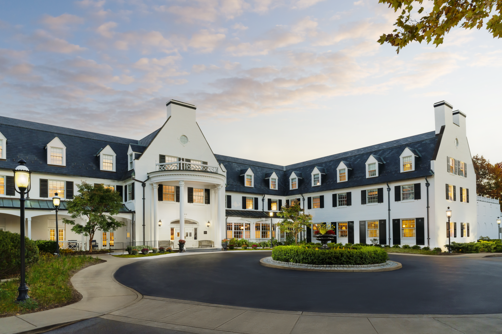 A large white hotel with a circular driveway, arched entrance, and symmetrical wings; well-kept landscaping and a sunny sky, inviting and upscale.