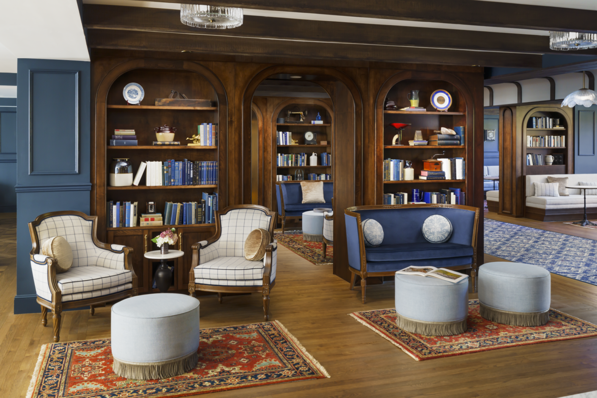 A classy library lounge with wooden arch shelves, blue seating, plaid chairs, and round ottomans on patterned rugs.