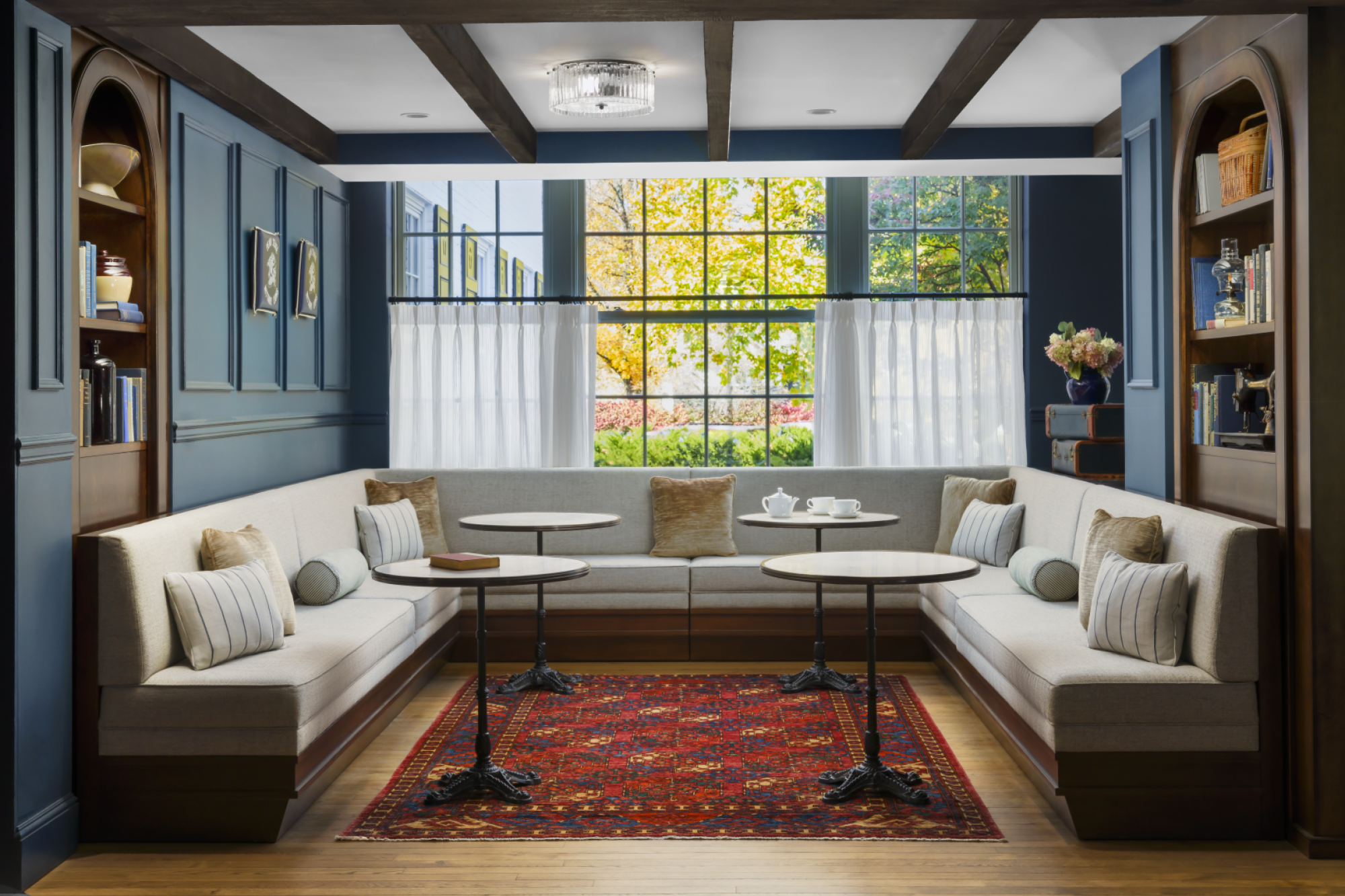 A cozy, built-in dining nook with blue paneled walls, white cushioned benches, and a central curved window. End tables, shelves with decor, a red patterned rug, and wooden ceiling beams. Ends with a period.