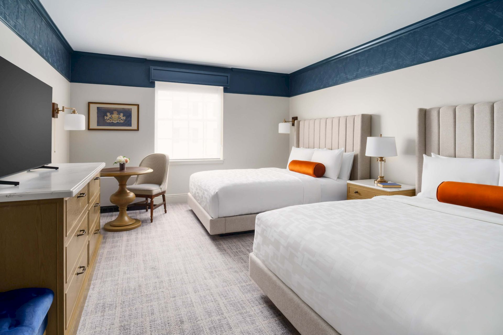 A bright hotel room with two queen beds, blue trim, white bedding, orange pillows, a desk area, and a large window.