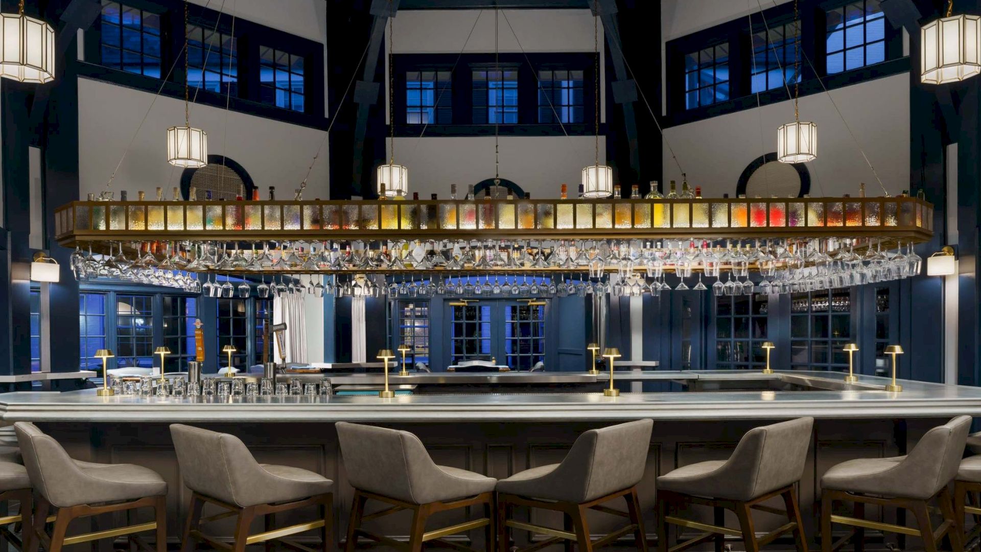 A stylish, dimly lit bar with a long counter, bar stools, and hanging lantern-style lights in a high-ceilinged room. 140 characters.
