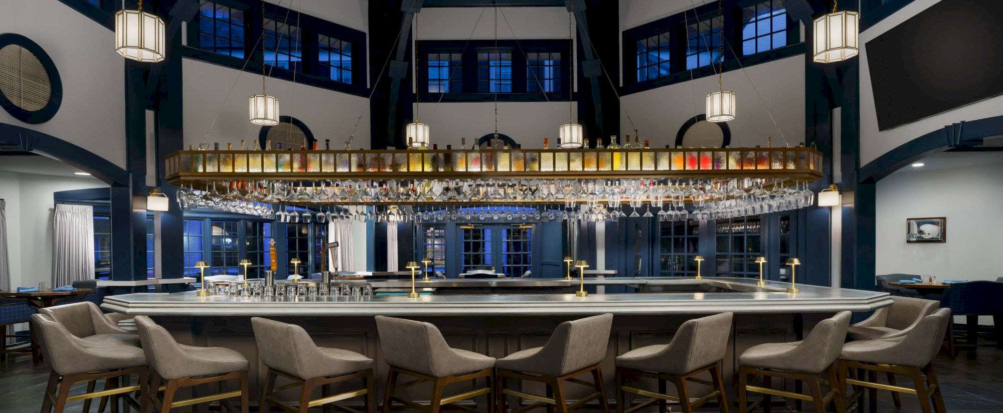 A stylish, dimly lit bar with a long counter, bar stools, and hanging lantern-style lights in a high-ceilinged room. 140 characters.