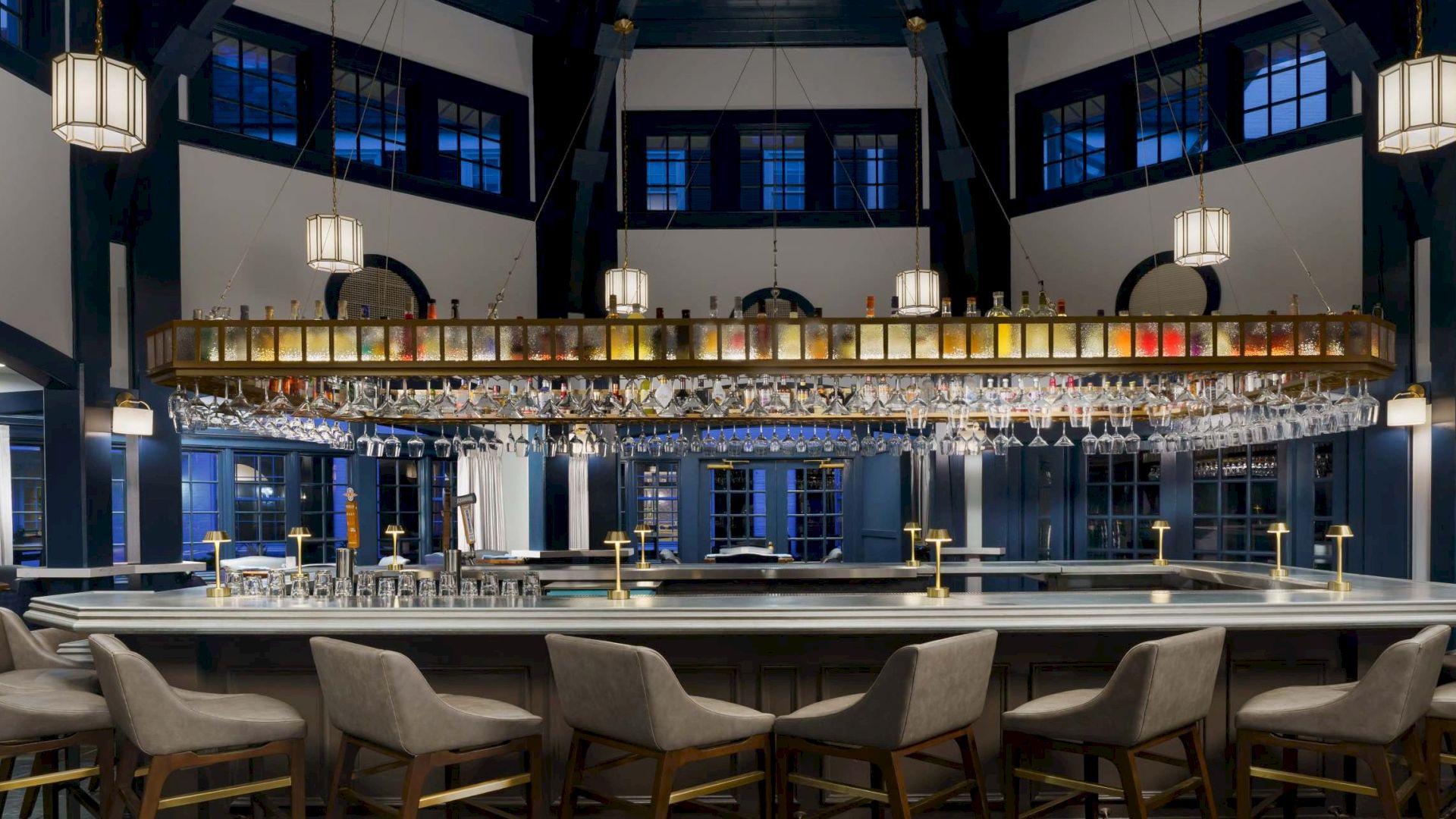 A stylish, dimly lit bar with a long counter, hanging lanterns, and blue-accented walls; modern chairs line the bar in a cozy setting.
