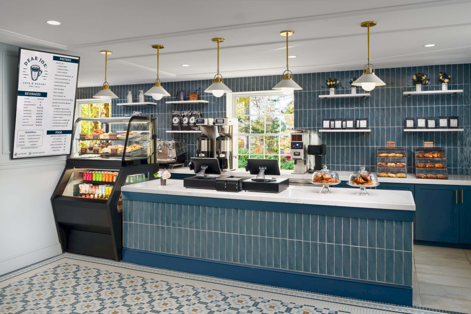 A cozy cafe counter with a blue tiled front, glass display case, pastry shelves, and hanging brass lamps; menu board on the wall and a sunny, airy vibe.