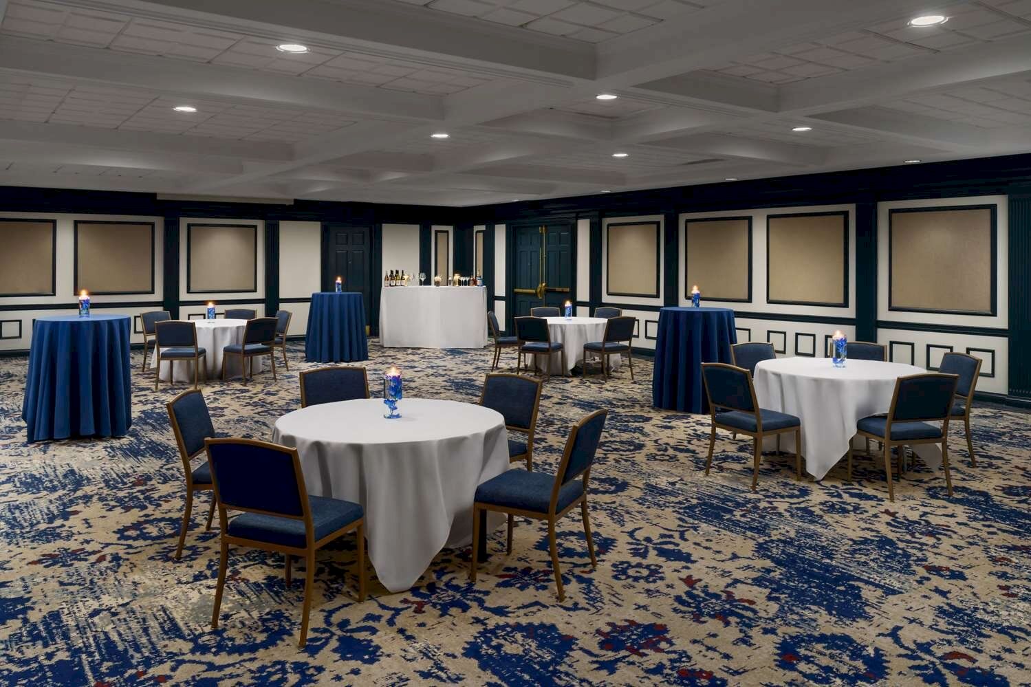 A banquet hall set with round tables covered in white cloths, blue chairs, and water bottles, ready for an event.