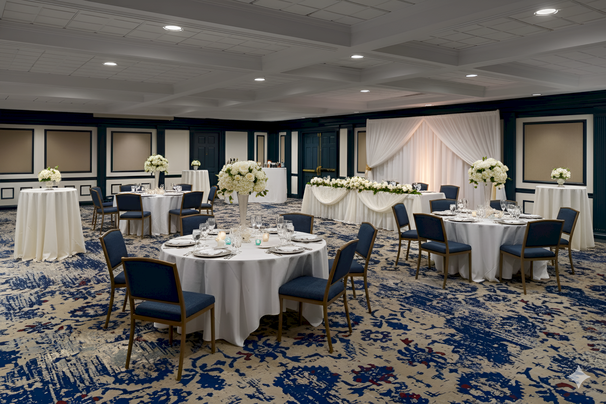 A banquet hall with round tables, white linens, elegant floral centerpieces, and a head table draped in white, ready for a wedding reception.