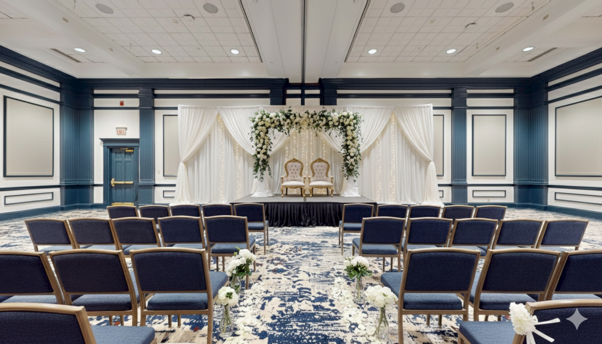 Elegant wedding ceremony setup: a floral arch backdrop with draped white fabric, two chairs on a stage, and rows of navy chairs facing forward.