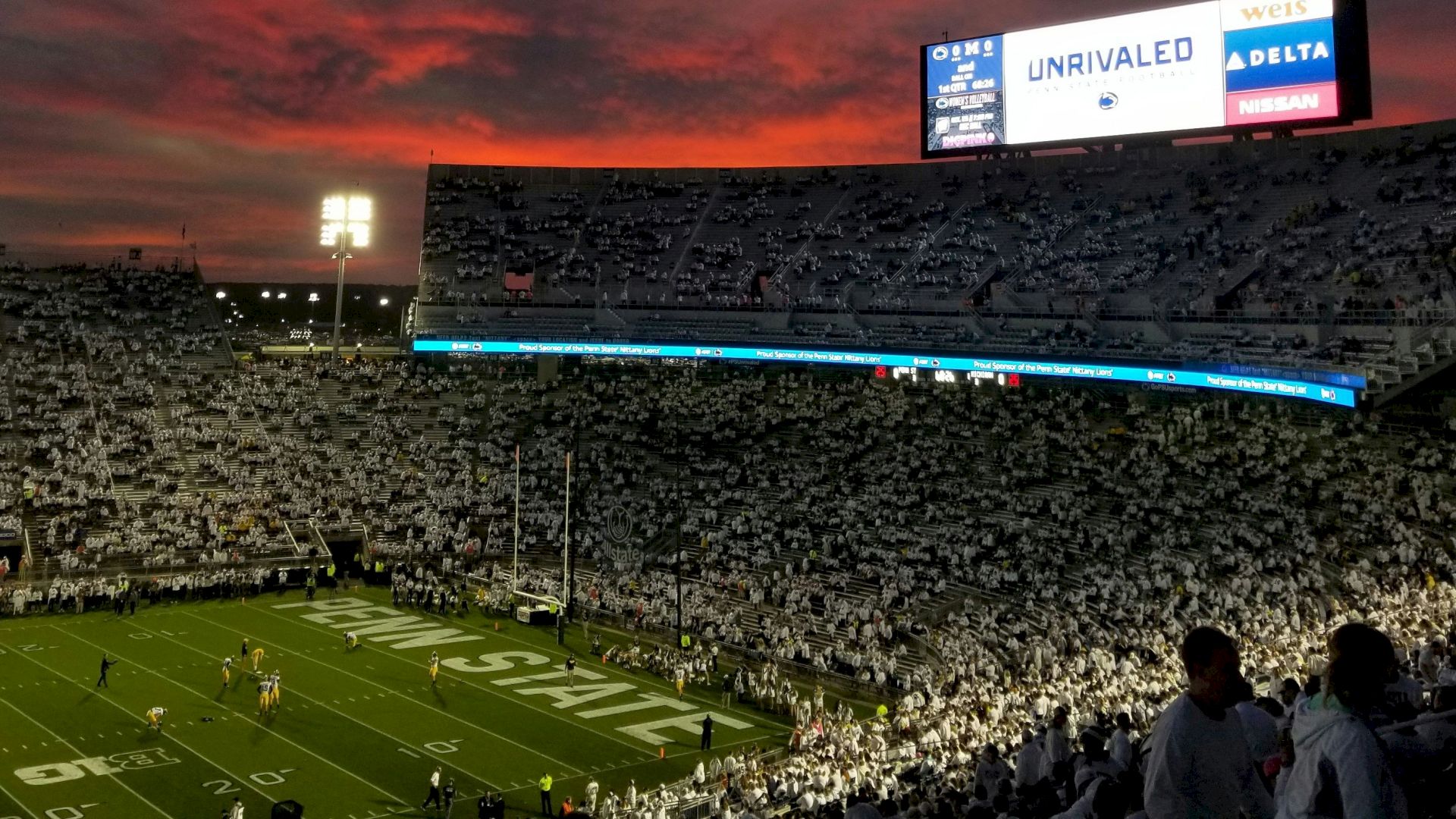 A packed football stadium at dusk, crimson sky, bright stadium lights, players on the field, and a large digital scoreboard glowing above the stands.