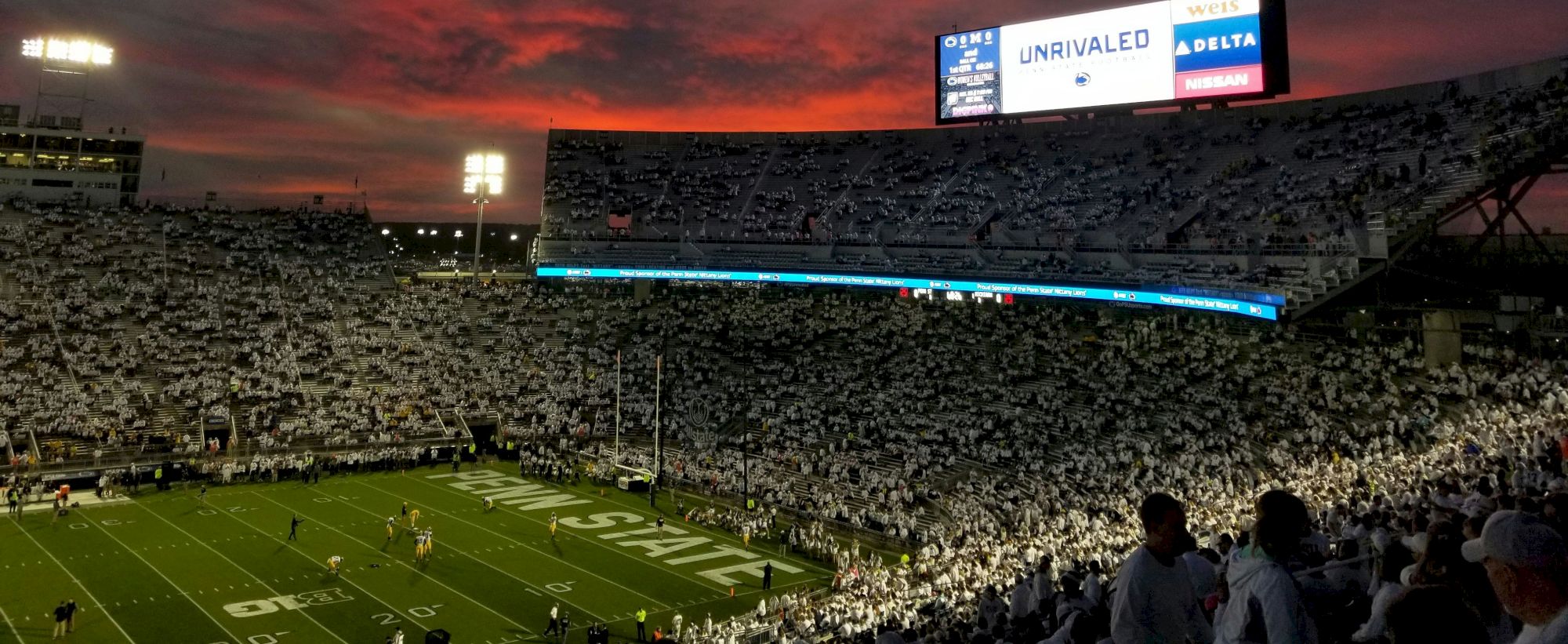 A packed football stadium at dusk, crimson sky, bright stadium lights, players on the field, and a large digital scoreboard glowing above the stands.