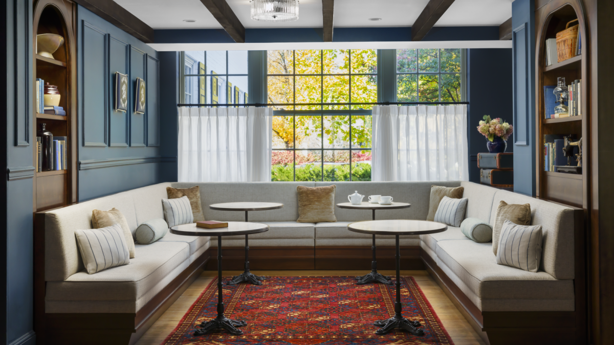 A cozy window-seat nook with built-in blue cabinetry, white cushions, round tables, a warm rug, and natural light streaming through a large pane.