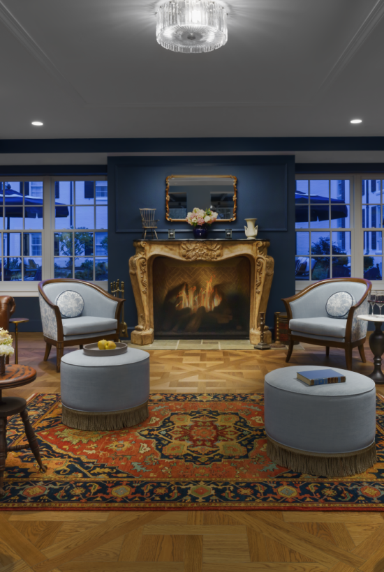 A luxurious living room with brown leather sofas, a grand ornate fireplace, blue walls, large windows, and round ottomans on a patterned rug, bright lighting.
