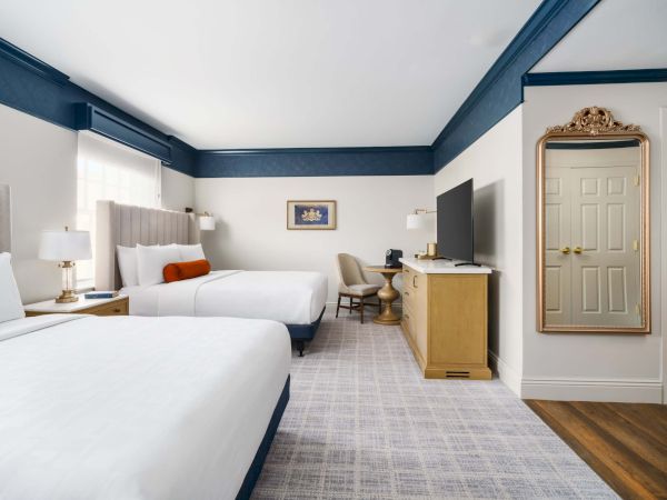 A hotel room with two queen beds, blue accents, a desk, TV, mirror, and a doorway; clean and modern, bright and airy.