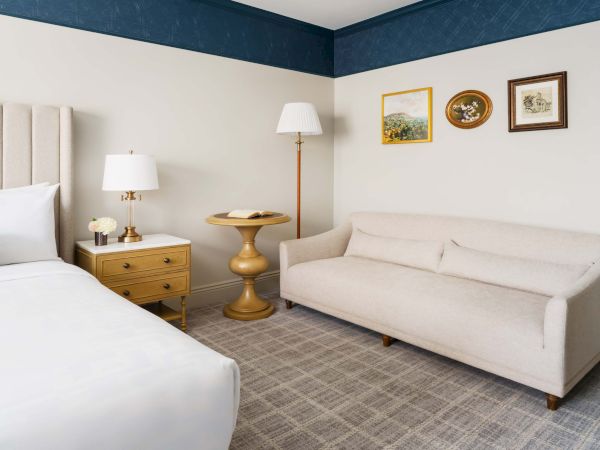 A bright hotel room with a white bed, beige headboard, nightstand, two lamps, a round gold side table, a beige sofa, framed art, and a blue accent wall, neat and modern.