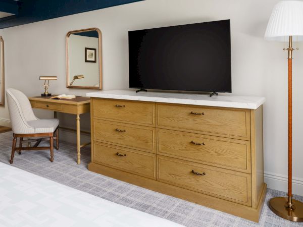 A wooden dresser with six drawers, a TV on top, a vanity desk with chair and mirror, and a tall floor lamp in a bright bedroom.
