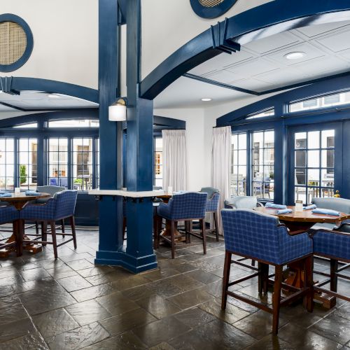 A stylish restaurant with blue and white decor, arched doorways, wicker chairs, dark wooden tables, and large windows letting in natural light.