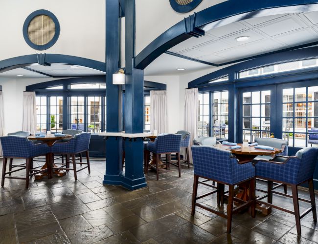 A stylish restaurant with blue and white decor, arched doorways, wicker chairs, dark wooden tables, and large windows letting in natural light.