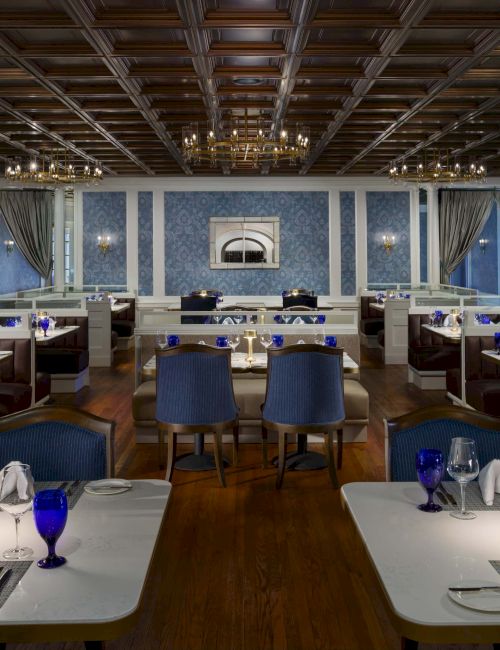 A stylish, blue-toned restaurant with white booths, wooden floors, blue glassware, and elegant chandeliers; intimate lighting and classical decor.