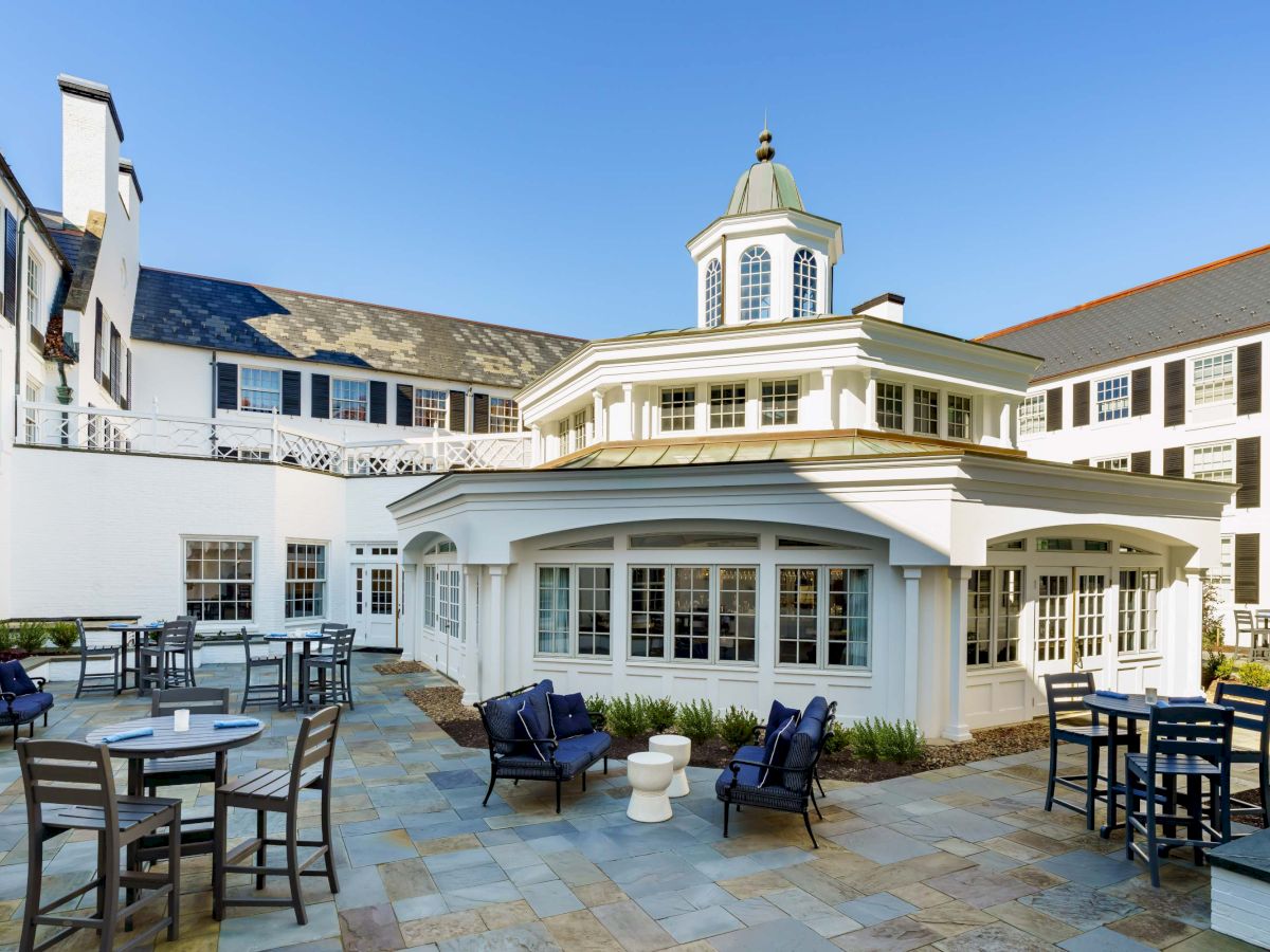 An elegant white hotel courtyard with a round glass-walled central lounge, outdoor seating, stone patio, and blue-sky backdrop, sunny and inviting.