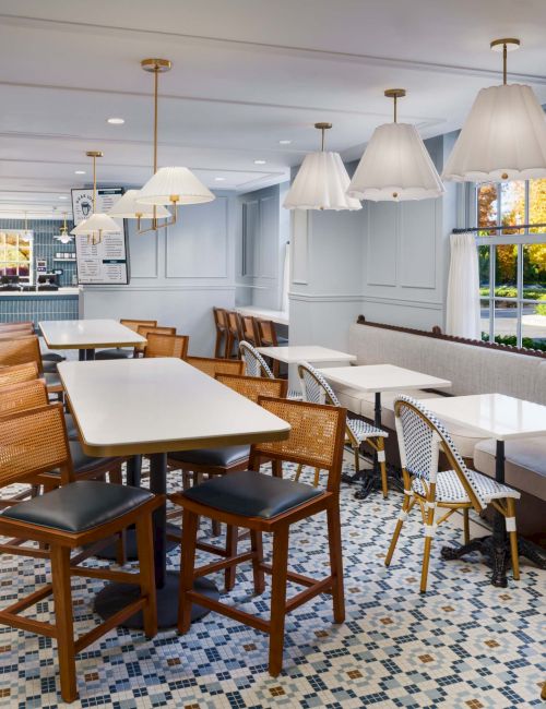 A bright, cozy dining room with mixed seating, long tables, vintage-style chairs, hanging lamps, patterned tiled floor, and a calm, welcoming vibe.