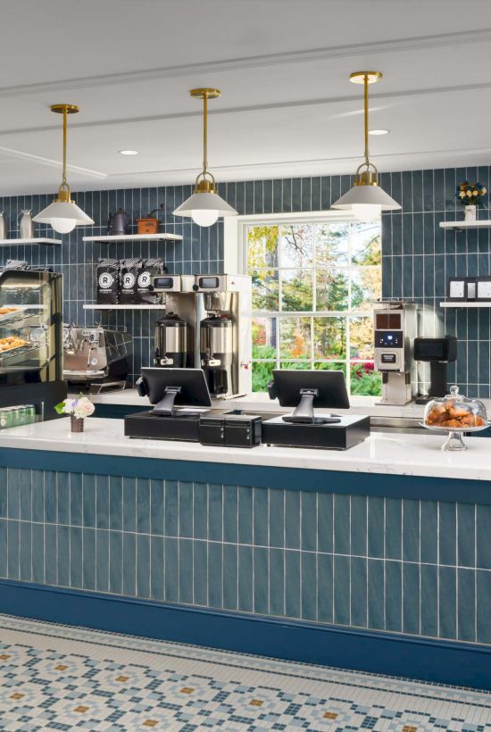 A bright cafe counter with blue tile front, white countertop, glass display case, pastries, coffee machines, shelves, and pendant lights.