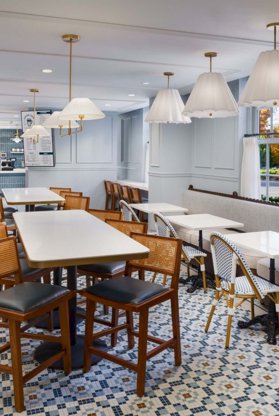 A bright, retro-style caf&eacute; dining area with wooden tables, mixed chairs, patterned tiled floor, and hanging white lamps.