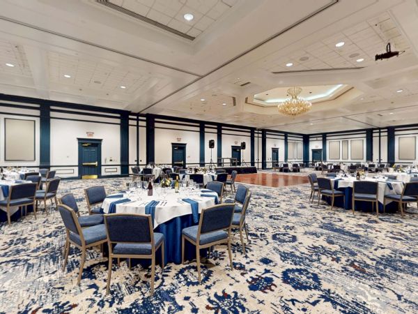A large banquet hall set for a formal event with round tables, blue chairs, white tablecloths, and elegant chandeliers in a spacious room.