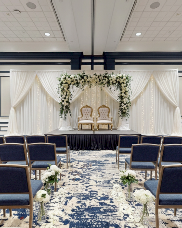 A bridal-style ceremony setup with a draped backdrop, floral arch, and two chairs on a small stage, facing rows of blue chairs.