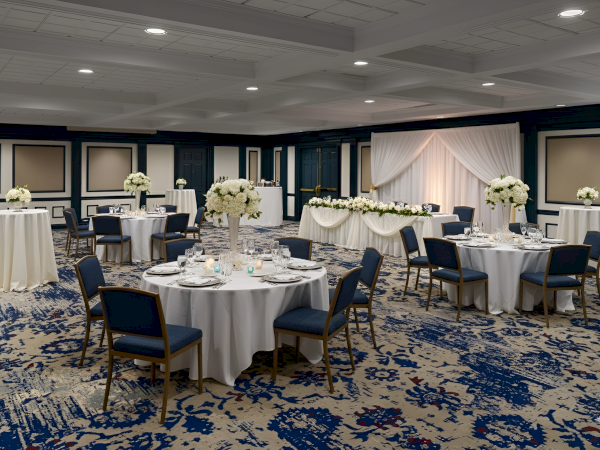 A banquet or wedding reception setup with round tables, white linens, blue chairs, and elegant white floral centerpieces in a decorated ballroom.