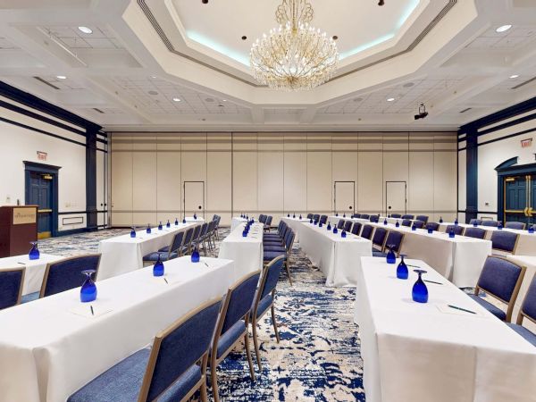 A large conference room with long white-clothed tables, blue chairs, blue bottles at each seat, chandeliers, and a carpeted floor in an elegant event setup.