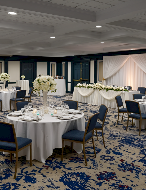 A banquet hall set for a formal event with round tables, white linens, blue chairs, elegant floral centerpieces, and a draped head table in the background.