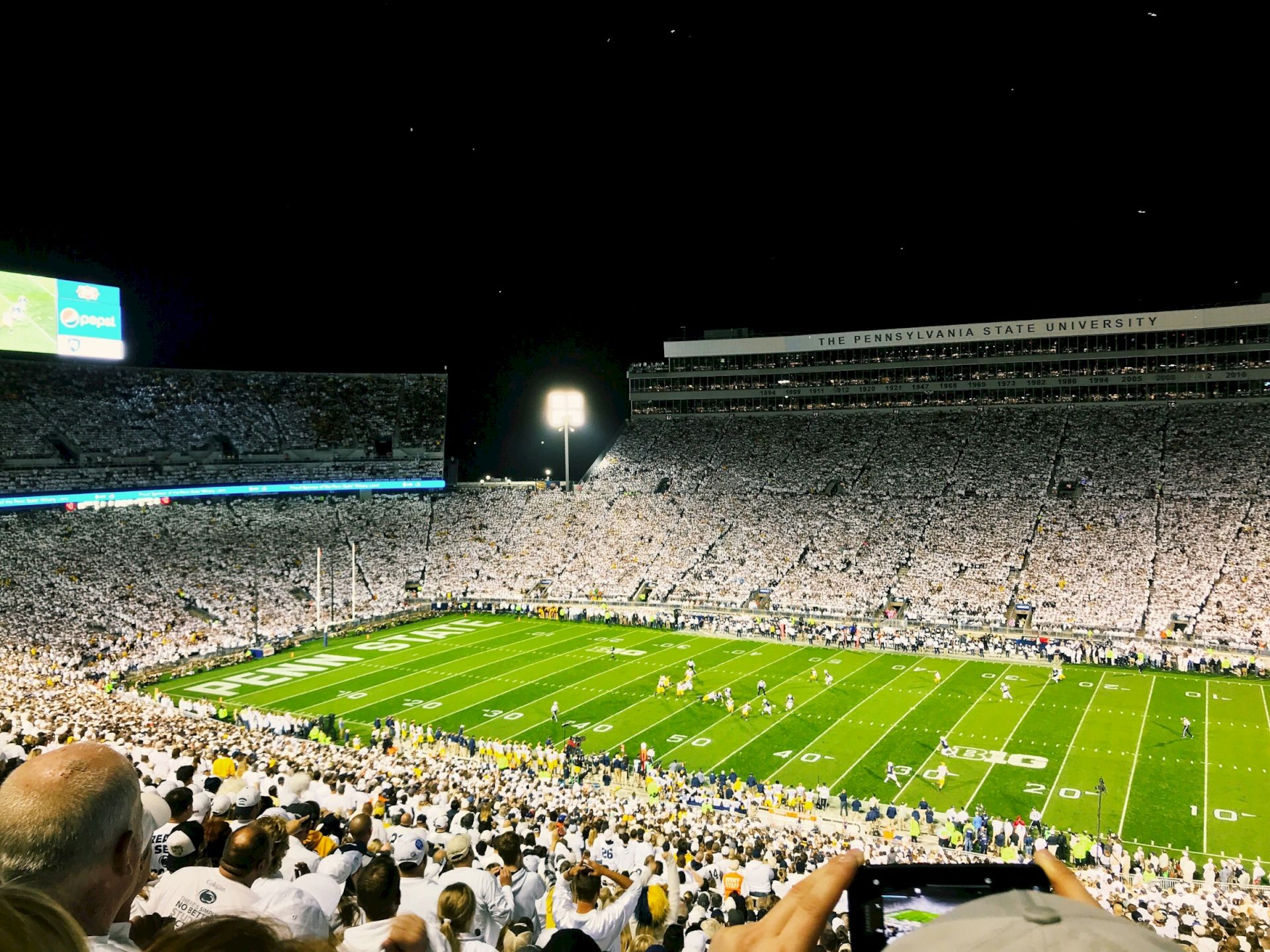 Packed Penn State football stadium at night, with a green field, bright screens, and thousands of fans cheering below.