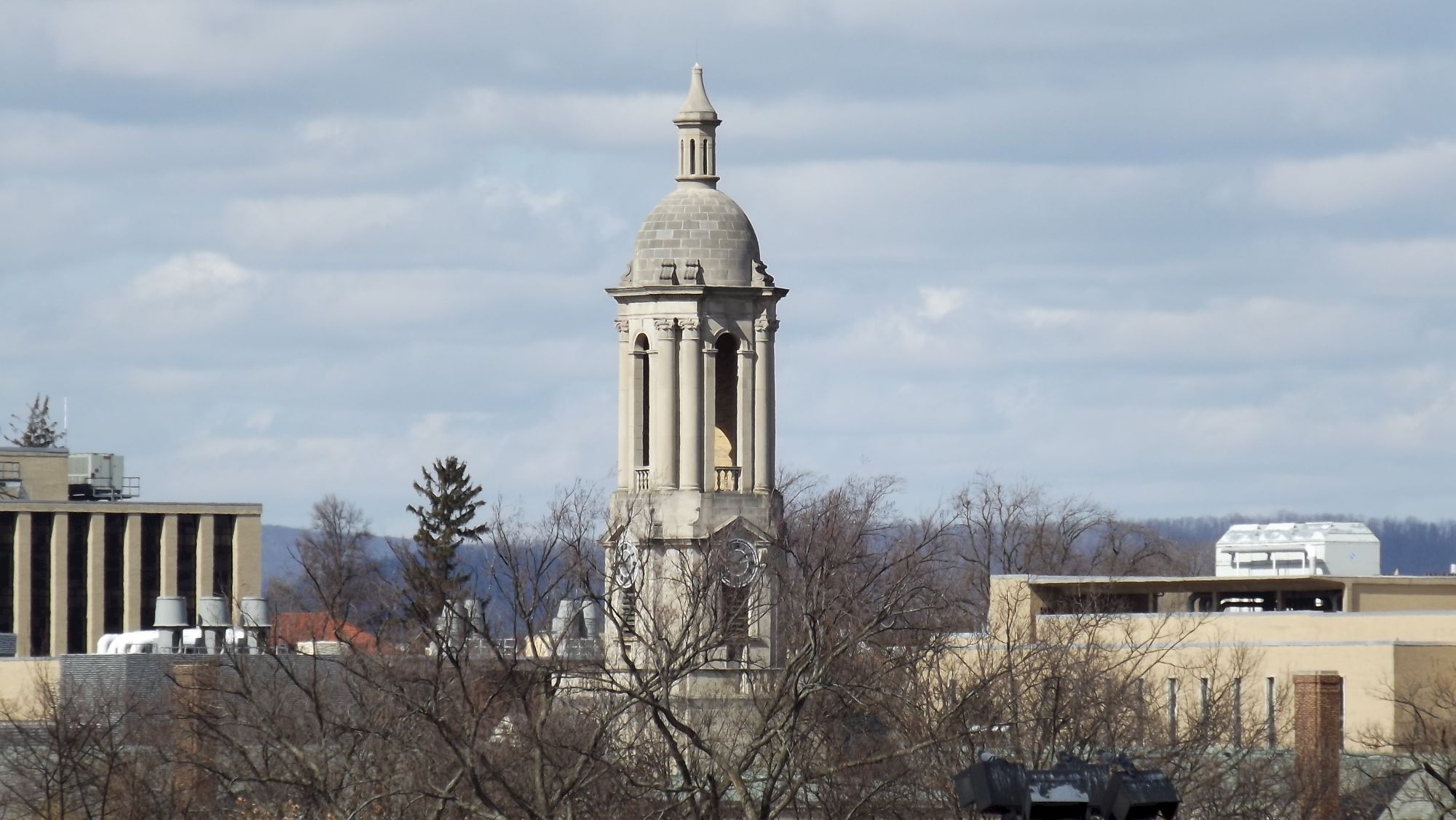 A city skyline with a tall domed clock tower rising above bare trees and surrounding buildings, under a partly cloudy sky.