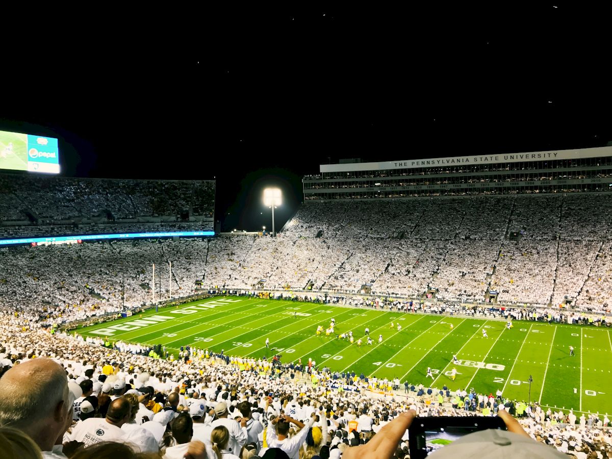 A packed football stadium at night, white-clad fans fill the seats, a bright green field glistens under stadium lights.