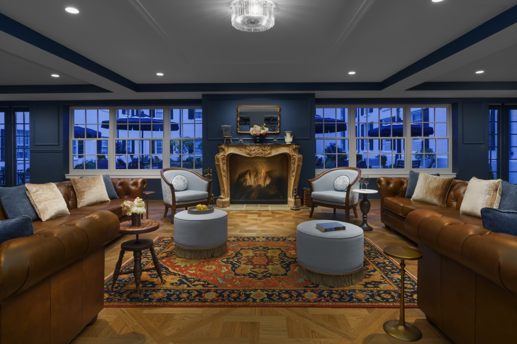 A luxurious lounge with brown leather sofas, blue-gray walls, a grand fireplace, and round ottomans around a patterned rug, under soft lighting.