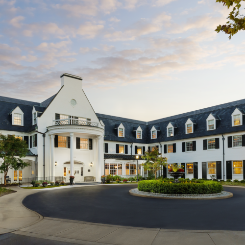 A row of modern townhouses with white and dark blue facades, gabled roofs, and a sunlit, tree-lined street.