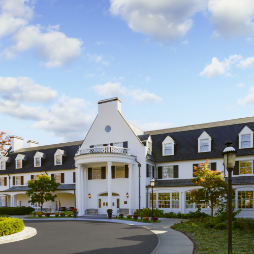 A sunny suburban street with neatly kept townhouses, green lawns, and a clear blue sky, cars parked along the curb, and trees in fall colors.