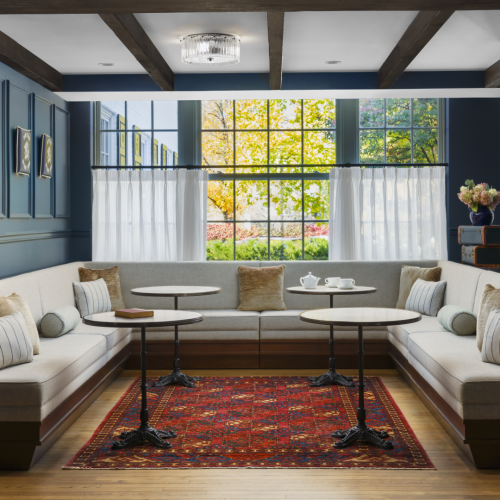 A cozy living room with a built-in bookshelf wall, a warm brick accent, large window with sunlight, and a plush sofa flanked by plants.