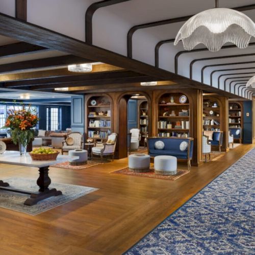 A spacious, elegant hotel lobby with wood beams, seating alcoves, a long carpeted corridor, and stylish pendant lights for a cozy, upscale vibe.