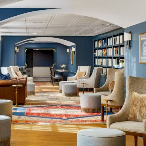 A cozy, blue-walled lounge with comfy chairs, ottomans, bookshelves, warm lighting, and a path of colorful rugs leading toward a TV area.