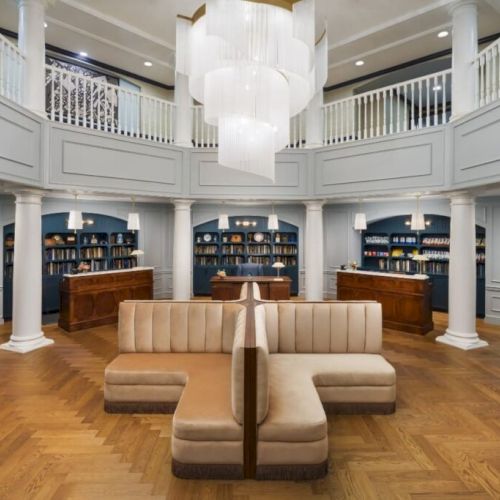 A spacious, elegant library or reading room with light wood flooring, classic white walls, arched bookcases, a central beige sofa, and a grand chandelier above.
