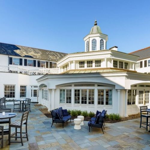 A bright, elegant hotel courtyard with white-clad buildings, outdoor seating, and a central glass-arched gazebo under a clear blue sky.